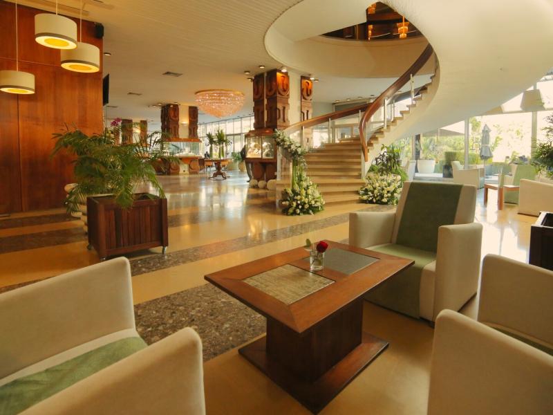 Stylish lobby lounge at Hotel Carlton showcases soft beige seating, rich wood finishes, and refreshing green accents beneath a sweeping modern staircase.
