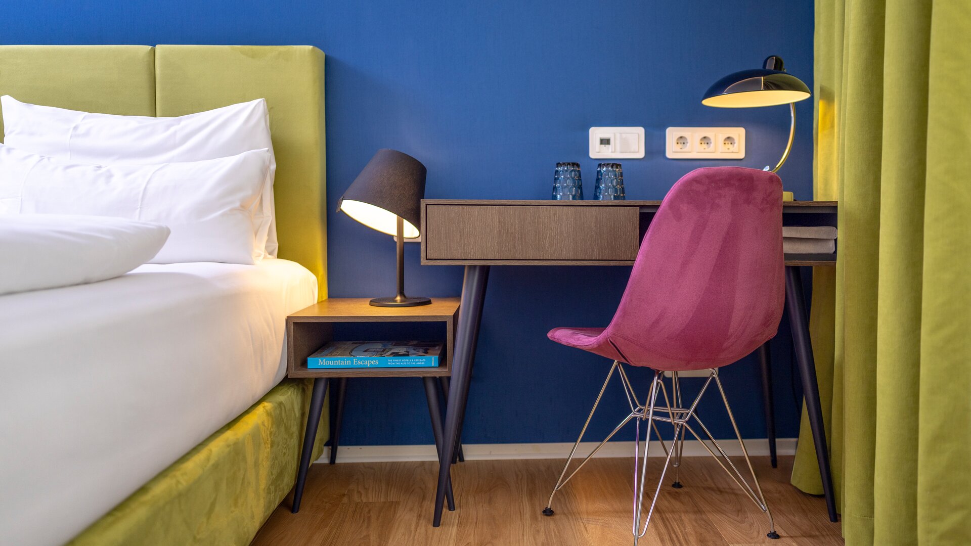Standard Guestroom Detail of Desk Area with a Pink Chair next to Bed with Green Frame and a Blue Accent Wall
