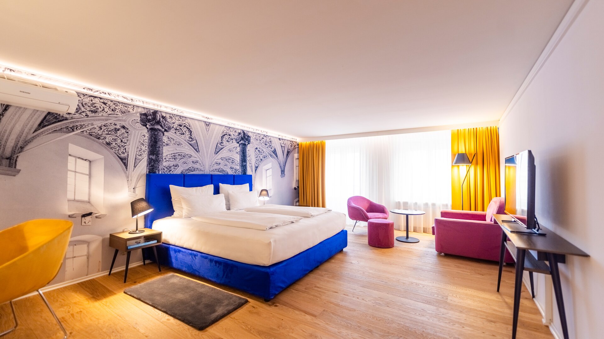 Junior Suite Overview with Blue, Pink, and Yellow Accents Decor, and with Backdrop of Black & White Mural Wall Paper of a Decorative Interior with Columns