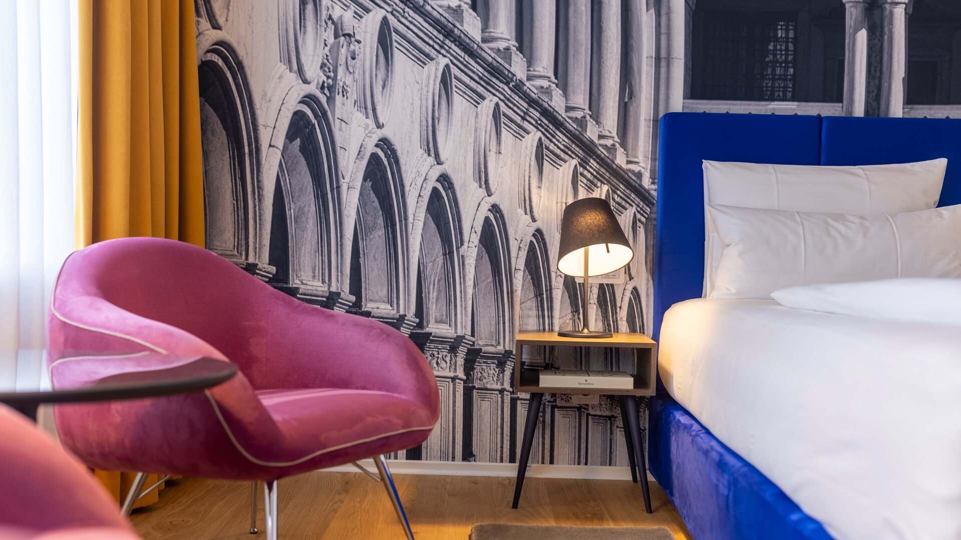 Junior Suite Detail View of Pink Chair, Nightstand and Bed with Accent Wall of Black & White Mural Wall Paper of Decorative Arches