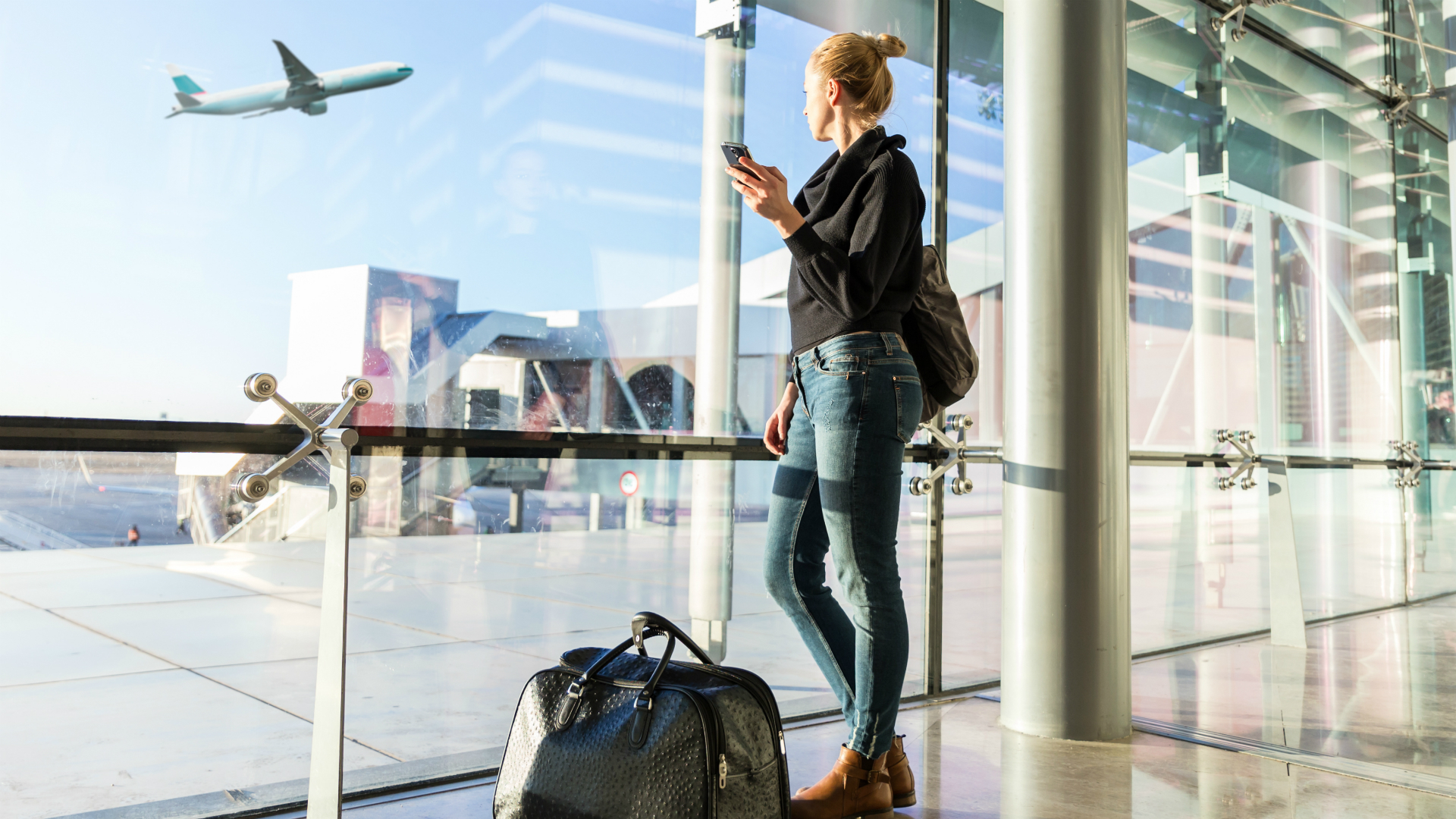 woman looking longingly out of airport window at a plan taking off into a clear blue sky