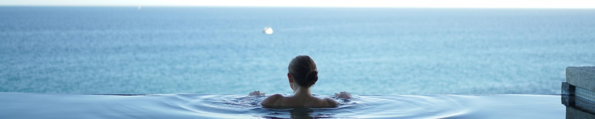 woman in a shaded infinity pool facing a blue ocean on a sunny day