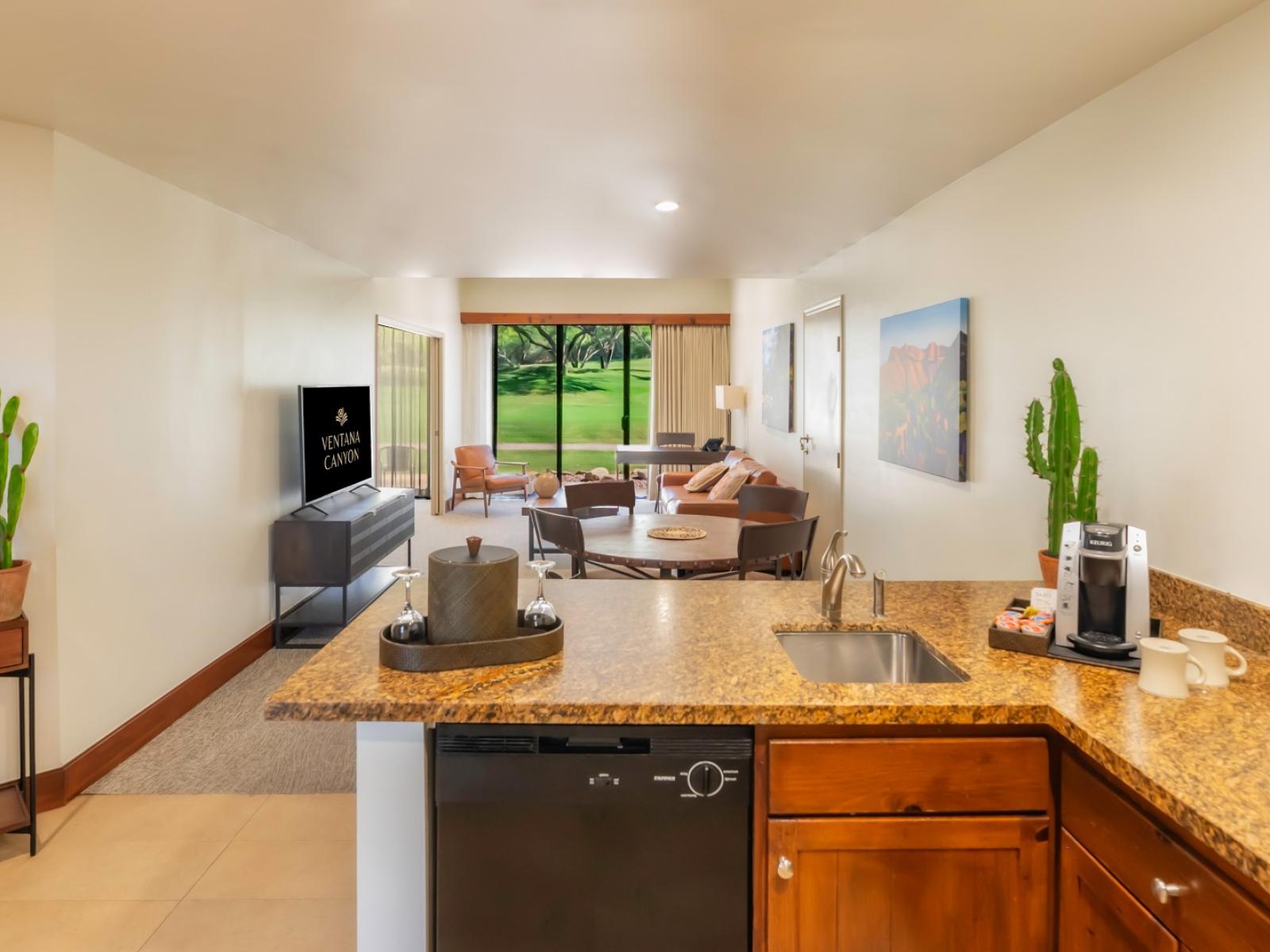 A view of the living room from the kitchenette, showing the dishwasher, counter and the couch and television in the living room beyond. 