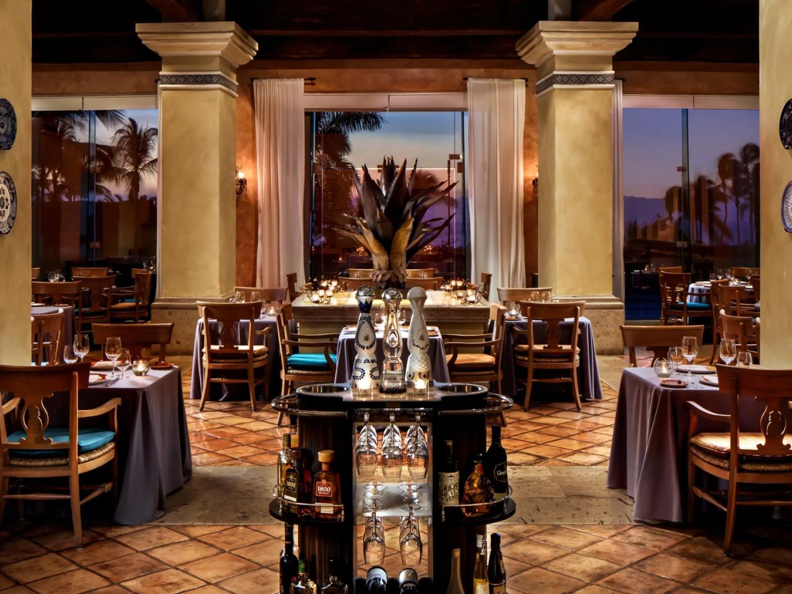 Grand Velas Riviera Nayarit Frida restaurant with set tables, warm lighting, decorative columns, and large windows showing palm trees outside.