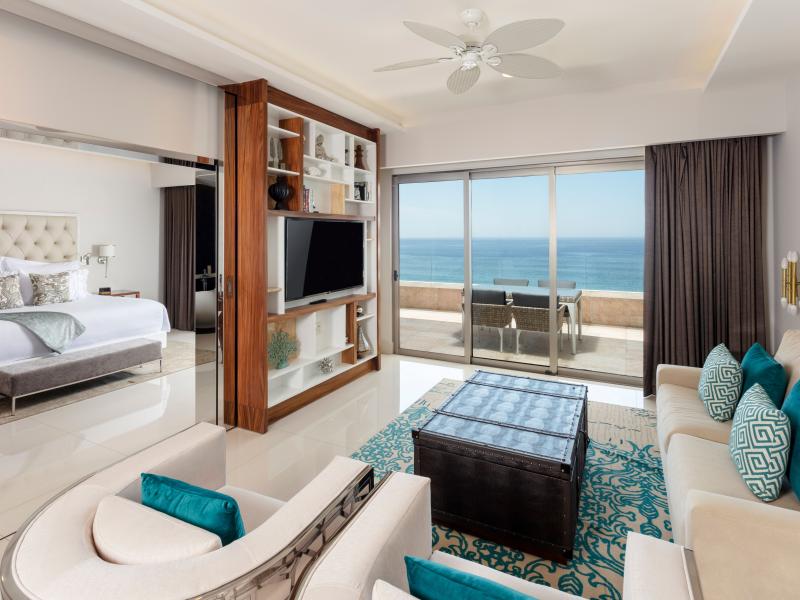 One Bedroom Suite features a cozy living area with teal accents, a separate bedroom, and floor-to-ceiling windows opening to a balcony with stunning ocean views.