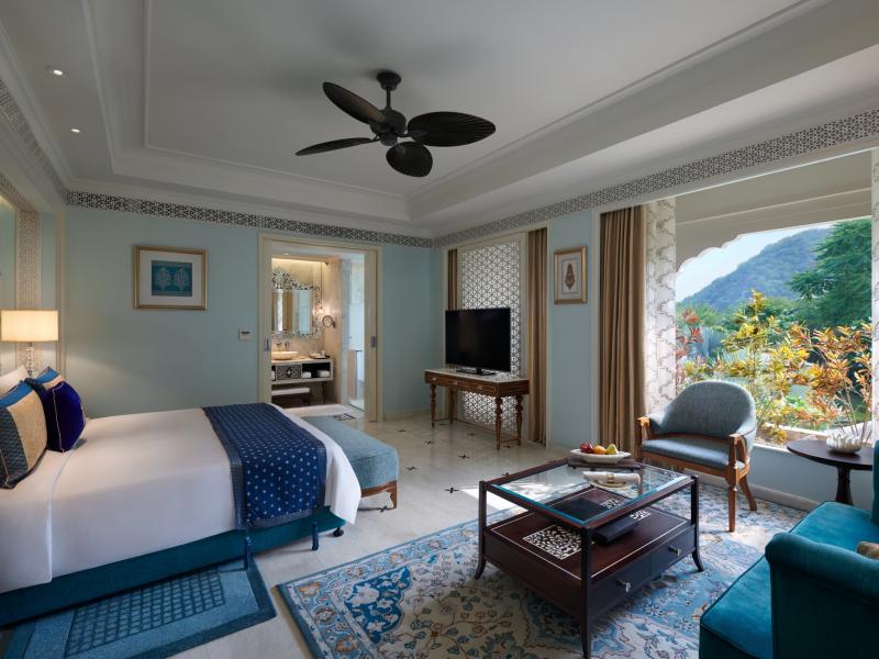 The serene bedroom of the Grand Villa at The Leela Palace Jaipur features a plush king bed, patterned rug, and cozy seating, accented with soft blue, ivory, and warm wood tones framed by garden views.