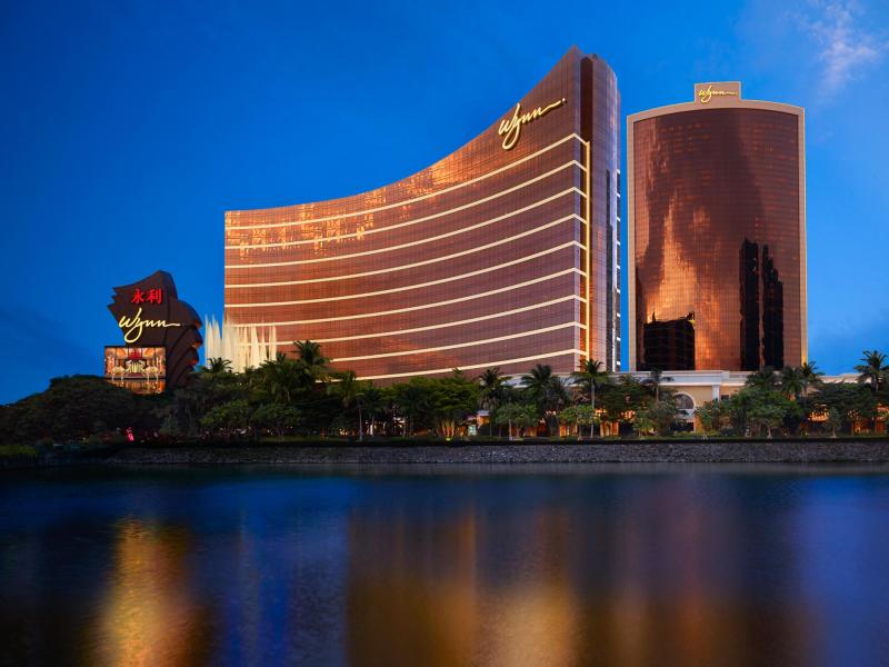 Reflecting over tranquil waterfront views, Wynn Macau and Encore striking bronze-glass façade glows against a deep sapphire sky, framed by lush emerald palms and golden illumination.