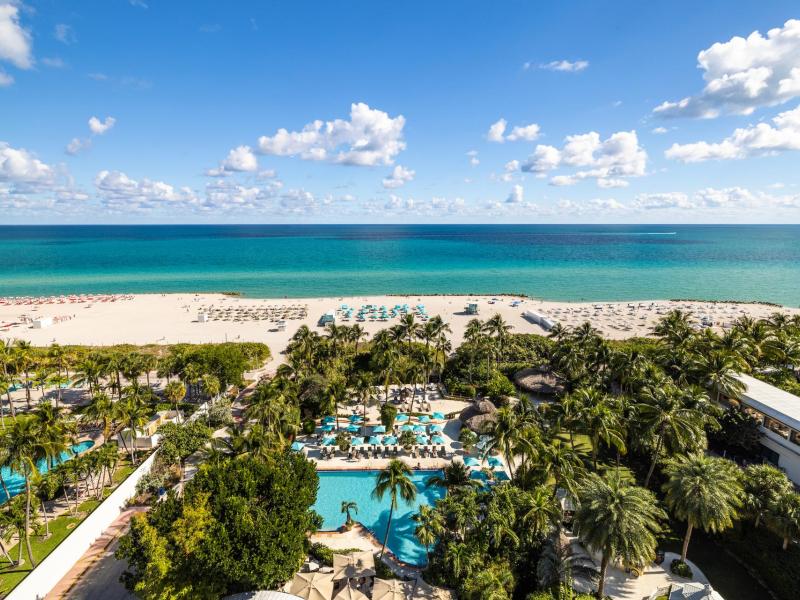 Aerial view of The Palms Hotel & Spa’s elevated beachfront oasis featuring lush tropical landscaping, a winding turquoise pool, and sandy shoreline accents highlighted by vibrant aqua blues, sandy whites, and fresh palm greens.