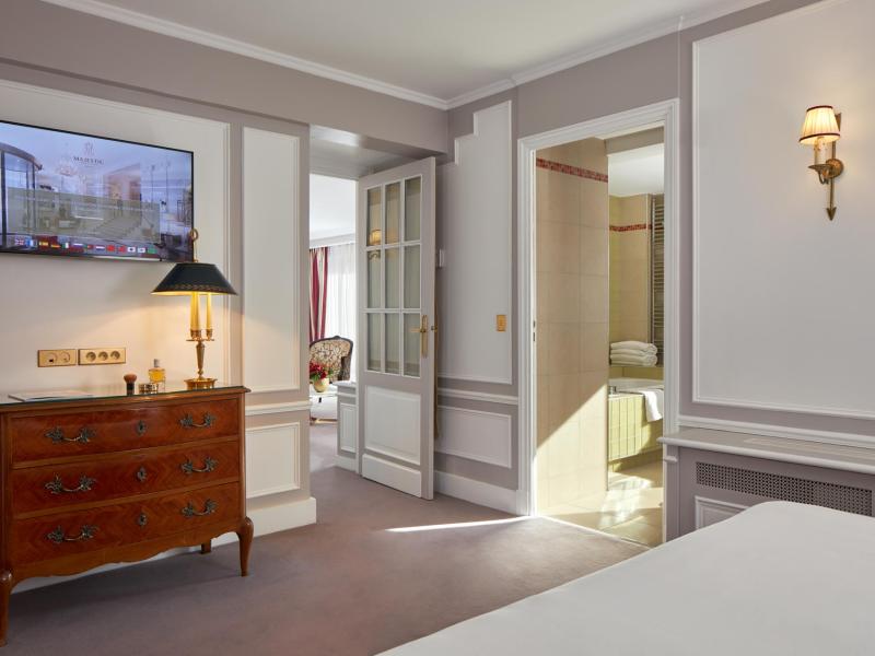 The Eiffel Tower Terrace Suite bedroom with paneled walls, antique wood furnishings, and an open view into a marble-lined bath, styled in warm gray, honey wood tones, and soft white.