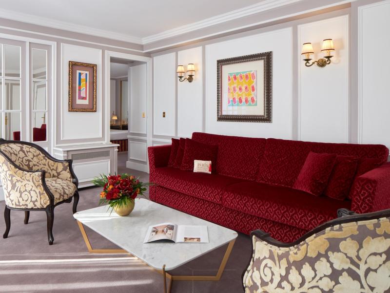 The Eiffel Tower Terrace Suite’s opulent living area showcasing plush upholstered seating, classic wall paneling, and curated artwork, richly accented in deep ruby red, soft taupe, and ivory.