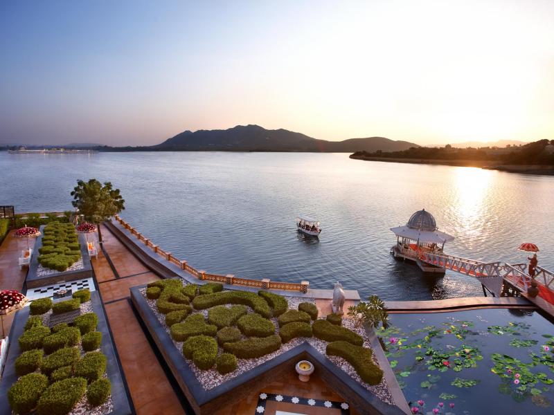 A tranquil sunset scene features a jetty sailing towards The Leela Palace Udaipur entrance dock with manicured gardens along the lake, accented with terracotta tones, vibrant greens, and gentle gold reflections.