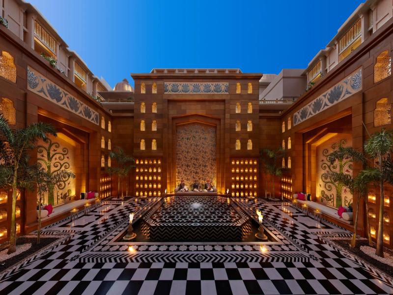 The Leela Palace Udaipur’s striking inner courtyard showcases a tiered fountain, ornate arches, and patterned stone flooring, accented with warm gold lighting and bold black‑and‑white details.