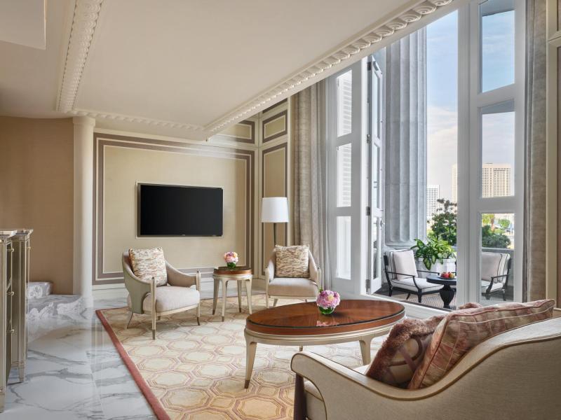 The Loft Suite’s bright living area showcases soaring windows, classic seating, and a polished marble floor accented by warm beige, soft pink, and gold tones.
