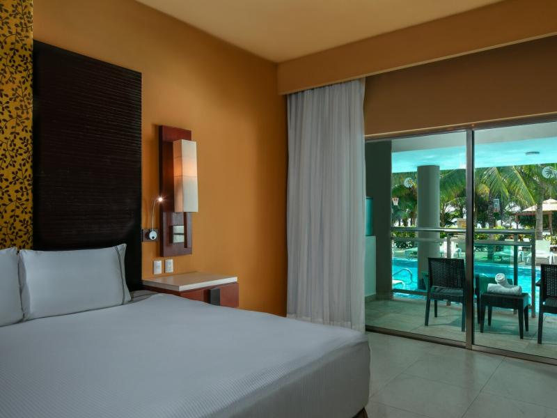 Oceanfront One Bedroom Presidential Suite with a large bed and a balcony overlooking a pool surrounded by palm trees.