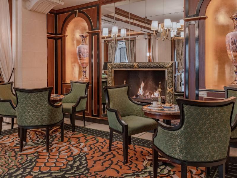 The lobby provides cozy seating around a glowing fireplace framed by rich mahogany panels, with green patterned chairs and golden highlights adding a touch of luxury.