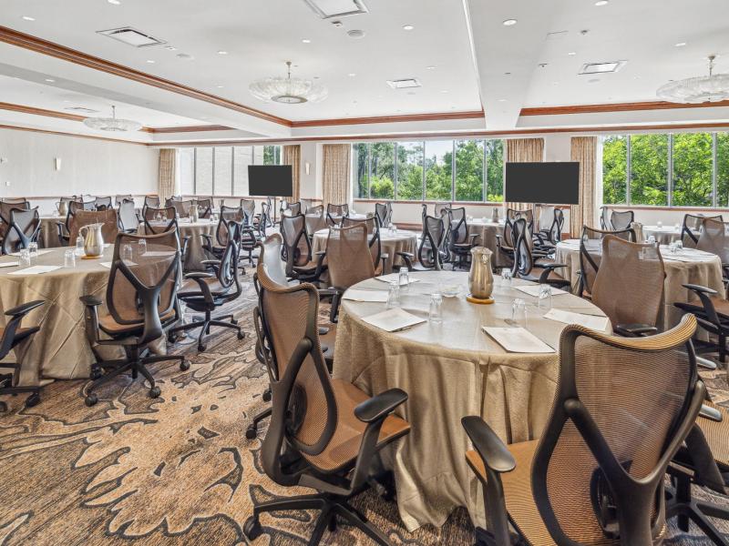 The Houstonian Hotel’s Aspen Room modern event space showcases round banquet tables, ergonomic rolling chairs, and expansive windows, styled in warm beige and taupe with rich wood accents and soft gray details.