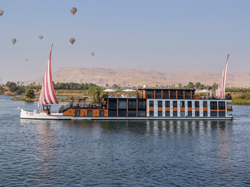 Luxurious river cruise vessel Mazaj Sia sails along serene waters under a sky dotted with colorful hot air balloons.