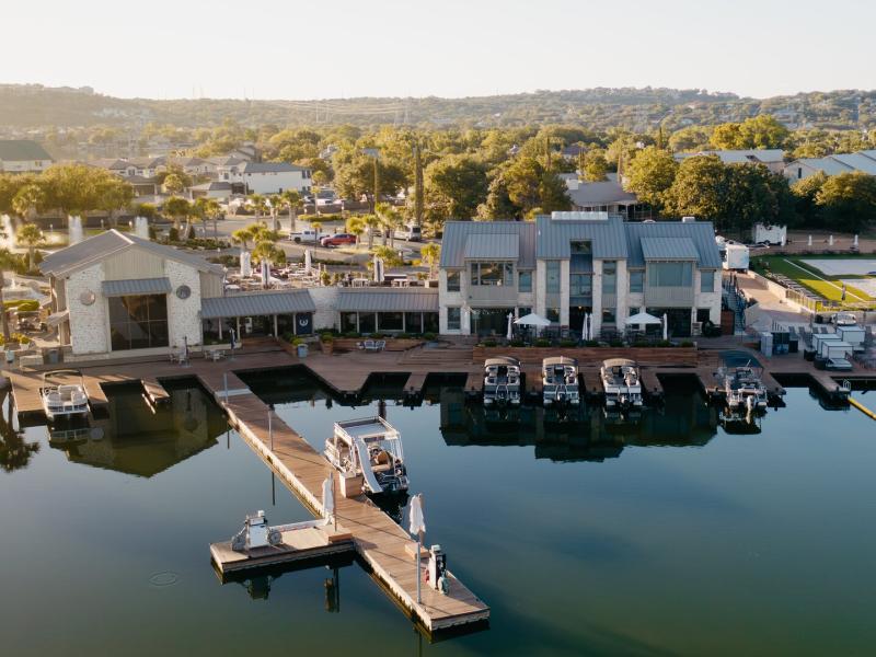 Horseshoe Bay Resort serene lakeside marina glistens under the sun, with sleek boats moored along wooden docks and a cluster of modern waterfront buildings framed by lush greenery and rolling hills in the distance.