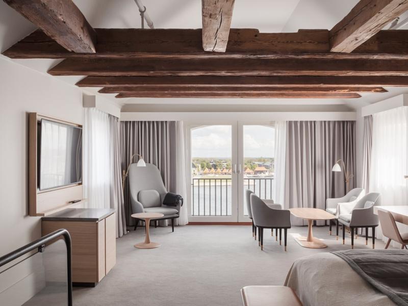A suite offering elegant comfort with rustic wooden beams, a king bed, multiple seating areas, and floor-to-ceiling windows framing stunning water views.