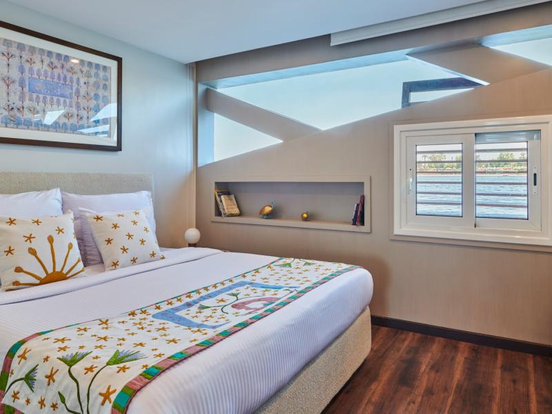 stylish cabin featuring a plush bed with vibrant accents, warm wood flooring, and unique windows framing serene river views.