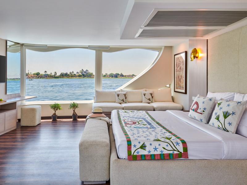 Spacious cabin suite with a plush king bed, elegant décor, and panoramic river views through floor-to-ceiling windows.