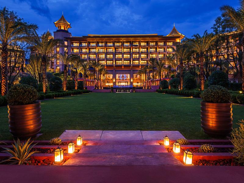 Exterior of hotel illuminated at night, framed by palm trees and lantern-lit pathways leading to its elegant entrance.