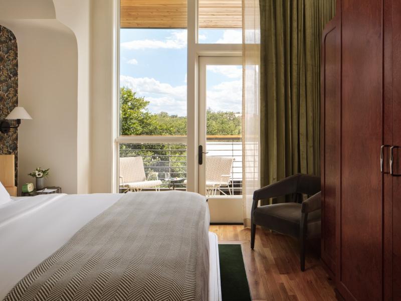 Cozy king guest room featuring a plush bed with crisp linens, a private balcony with outdoor seating, rich wood furnishings, and floor-to-ceiling windows framing serene views.