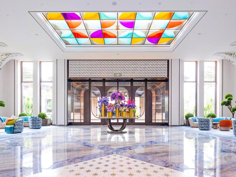 Bright and spacious lobby with colorful geometric ceiling panels, elegant floral arrangements, and modern seating areas surrounded by lush greenery.