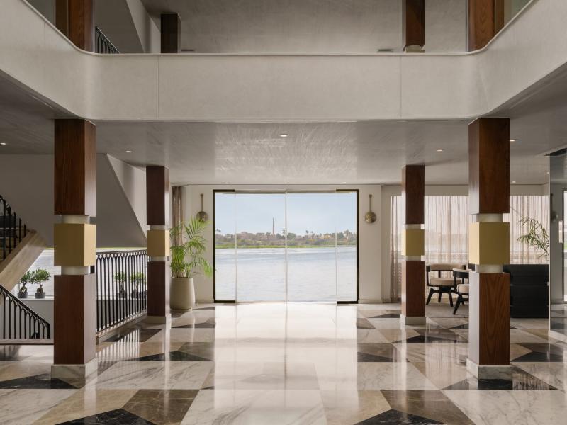 Step into a grand, light-filled lobby featuring striking geometric marble floors, sleek wood accents, and panoramic glass doors framing stunning waterfront views.