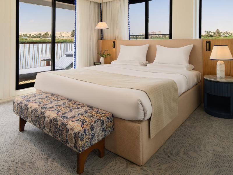Suite or cabin featuring a plush king bed, patterned upholstered bench, soft neutral tones, bedside lamps, and floor-to-ceiling sliding doors opening to a private balcony.