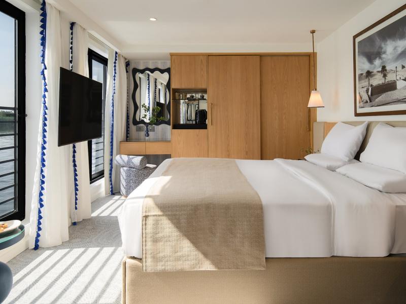 Suite or cabin bedroom with king bed, sleek wooden wardrobe, wall-mounted TV, framed artwork, and expansive windows offering uninterrupted views of the tranquil river.