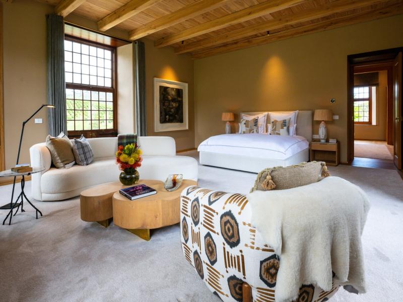 A residence's serene bedroom with a king bed, soft beige walls, a plush cream sofa, and a patterned armchair in earthy tones, all accented by warm oak ceiling beams and large grid windows framing lush garden views.