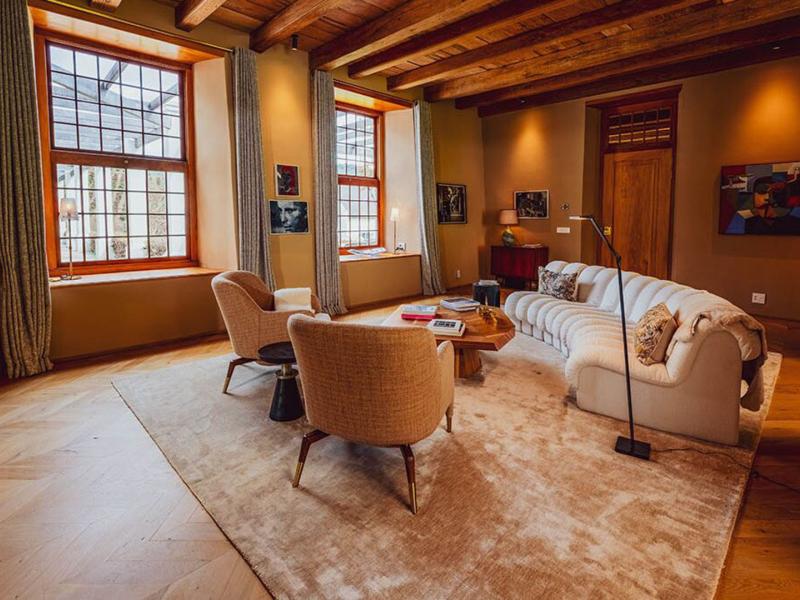 A residence's refined elegant lounge featuring rich wooden ceiling beams, taupe walls, a cream tufted sofa, and sand-colored armchairs atop a velvety beige rug, illuminated by natural light through tall grid windows