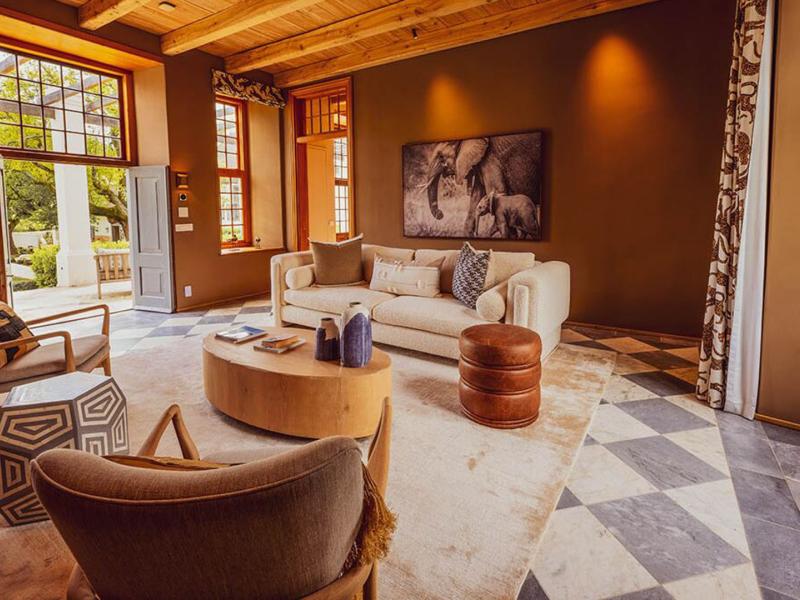A residence's sophisticated sitting room with mocha-toned walls, a cream sofa, caramel leather ottomans, and modern accent chairs in muted gray, complemented by black-and-white patterned curtains and sunlit garden views.