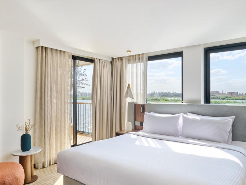 Sunlit suite bedroom with a plush king bed, floor-to-ceiling windows, sheer curtains, and a private balcony offering sweeping river views.