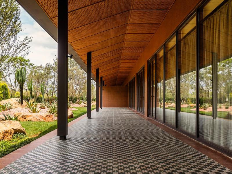 Covered outdoor corridor with patterned tile flooring, glass walls, and views of a landscaped garden featuring rocks and greenery.