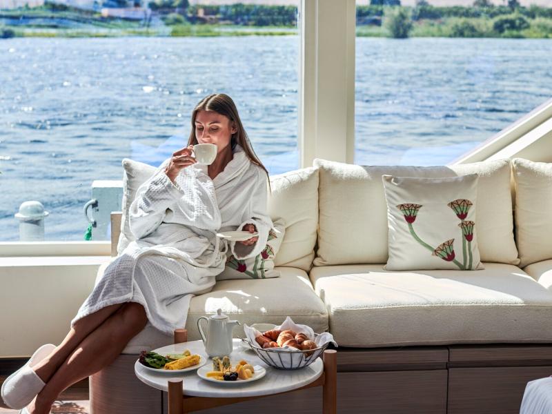 A relaxing moment in a plush cabin suite featuring a cozy sofa, elegant décor, and panoramic river views complemented by a gourmet breakfast spread.