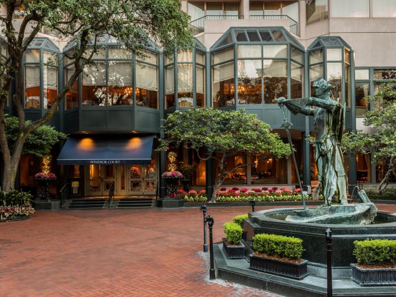 The Windsor Court charming entrance courtyard featuring a striking bronze fountain surrounded by manicured greenery and vibrant floral accents, framed by it’s elegant glass windows and a deep navy awning for a sophisticated entrance