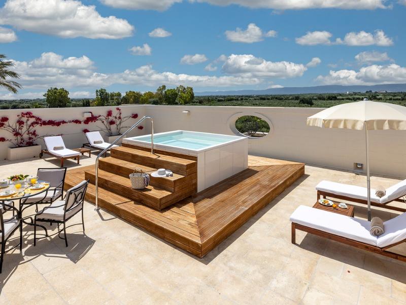 Terrace Suite chic rooftop featuring a sleek wooden-deck jacuzzi and elegant white loungers, accented by vibrant pink bougainvillea and soft beige stone under a bright blue sky.
