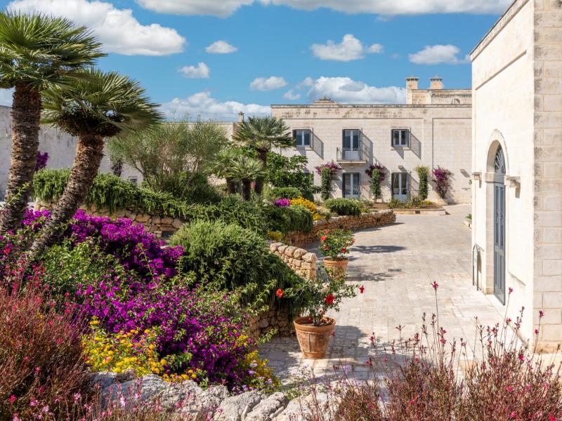 Masseria San Domenico elegant stone architecture surrounded by vivid purple and yellow blooms, with palm accents and a serene cobblestone courtyard under a crisp blue sky.