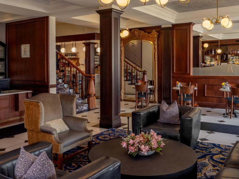 The Adelphi Hotel lobby featuring rich wood paneling and plush leather seating with soft gray and taupe accents, complemented by a chic bar and warm globe lighting for a sophisticated ambiance.
