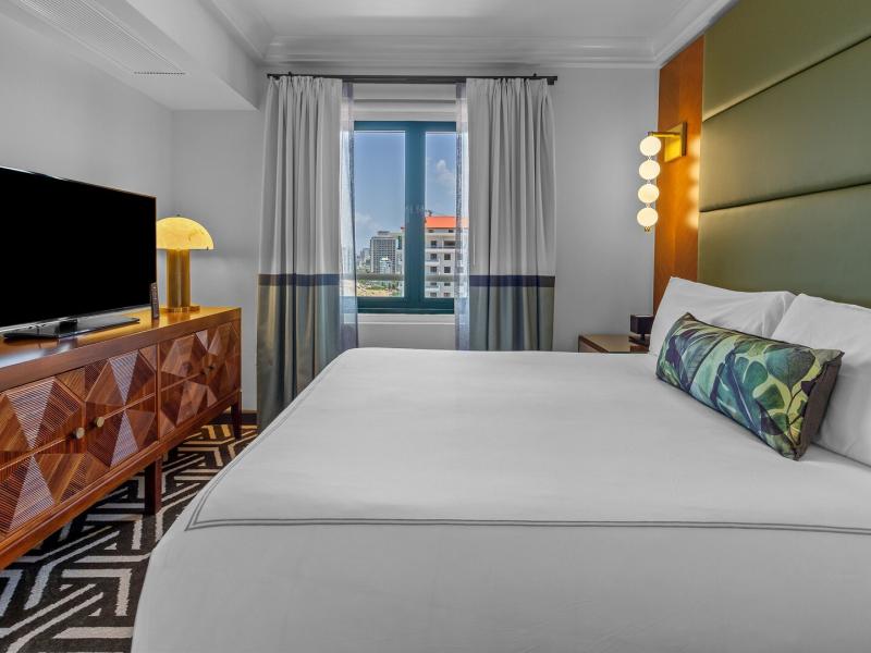 Commodore Oceanfront Suite serene bedroom featuring a plush king bed, geometric-patterned rug, and a large window framing vibrant city views of San Juan.