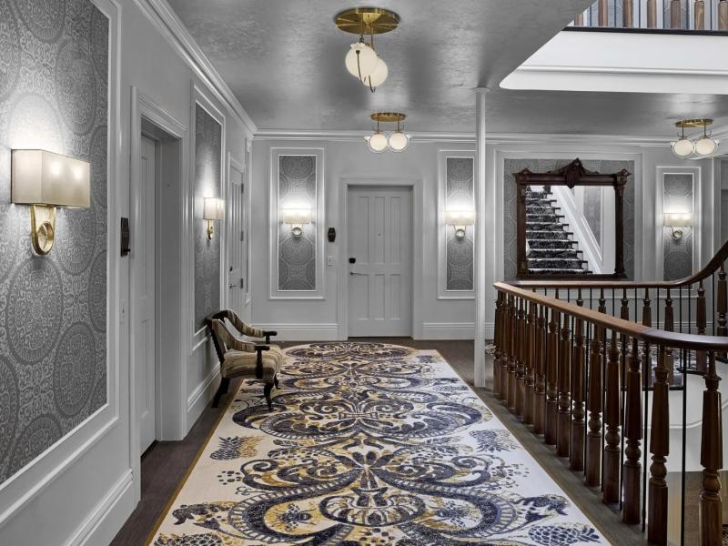 Corridor to guest accommodations at The Adelphi Hotel, highlighting intricate patterned carpeting in navy and gold against crisp gray walls, paired with classic wooden railings and brass light fixtures for timeless charm.
