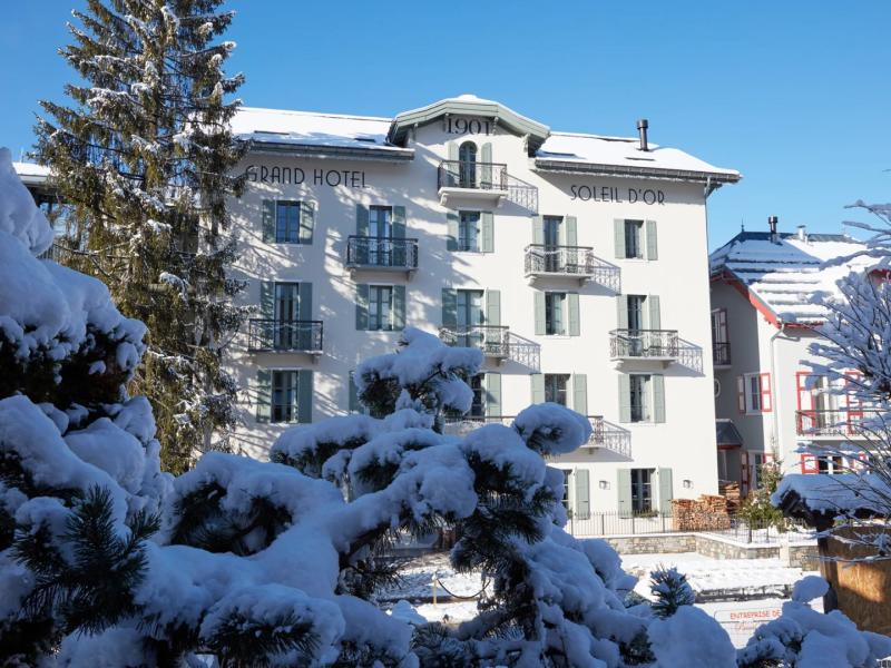 Grand Hôtel Soleil d’Or exterior showcasing a grand white façade with elegant balconies, framed by snow-covered pine trees and accented with soft gray and deep green tones.