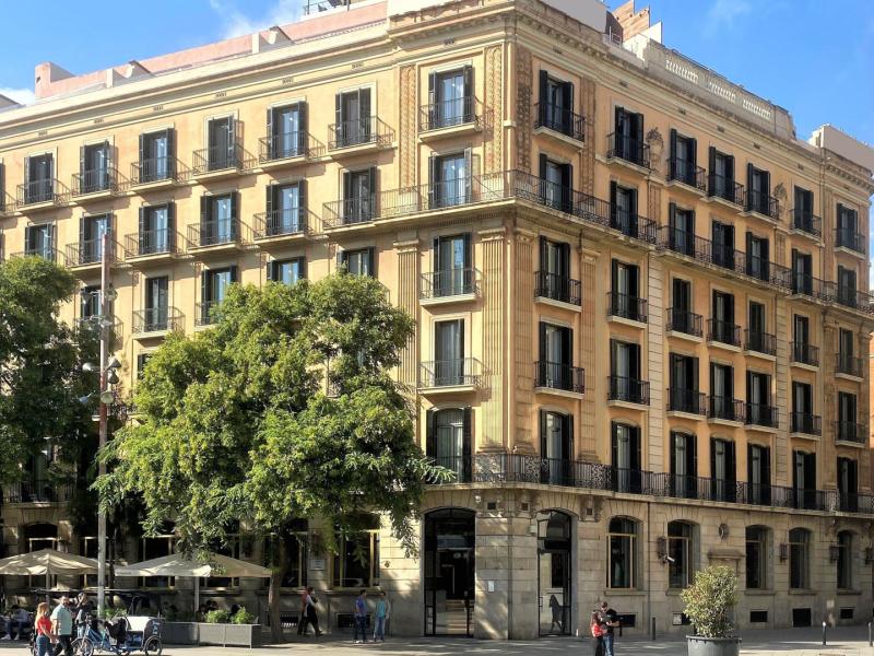 Lamaro Hotel showcasing it’s grand historic façade with elegant black-framed windows and warm beige stone accents, complemented by lush green trees and inviting outdoor seating.