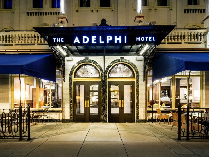 The Adelphi Hotel front entrance showcasing elegant arched double doors framed by warm lighting and bold black signage, accented with deep blue awnings and golden tones for a grand welcome.