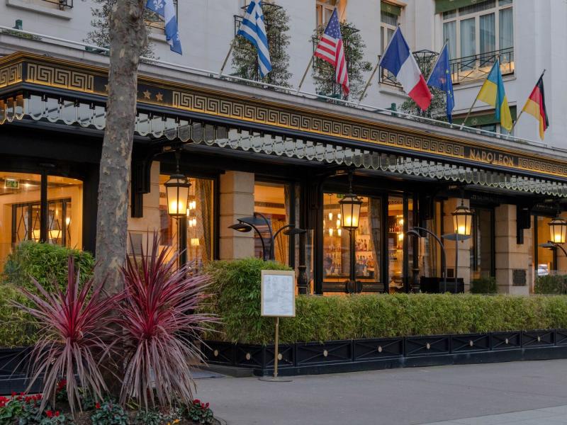 Hotel Napoleon Paris, showcasing a grand entrance with ornate black-and-gold detailing, vibrant red plants, and colorful international flags for a sophisticated welcome.