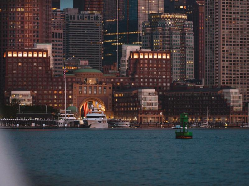 A stunning waterfront view of Boston Harbor Hotel with it's brick architecture and sleek modern skyscrapers in the background, accented by warm amber lights and deep blue waters.