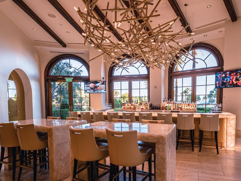 The elegant lobby bar with a gleaming marble counter and plush tan seating, highlighted by dramatic sculptural chandeliers and rich wood-framed arched windows.