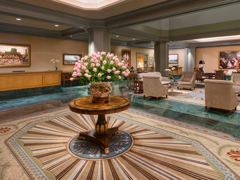 The Windsor Court spacious lobby featuring a striking patterned rug and polished wood centerpiece adorned with soft pink blooms, complemented by green marble accents and neutral-toned seating.